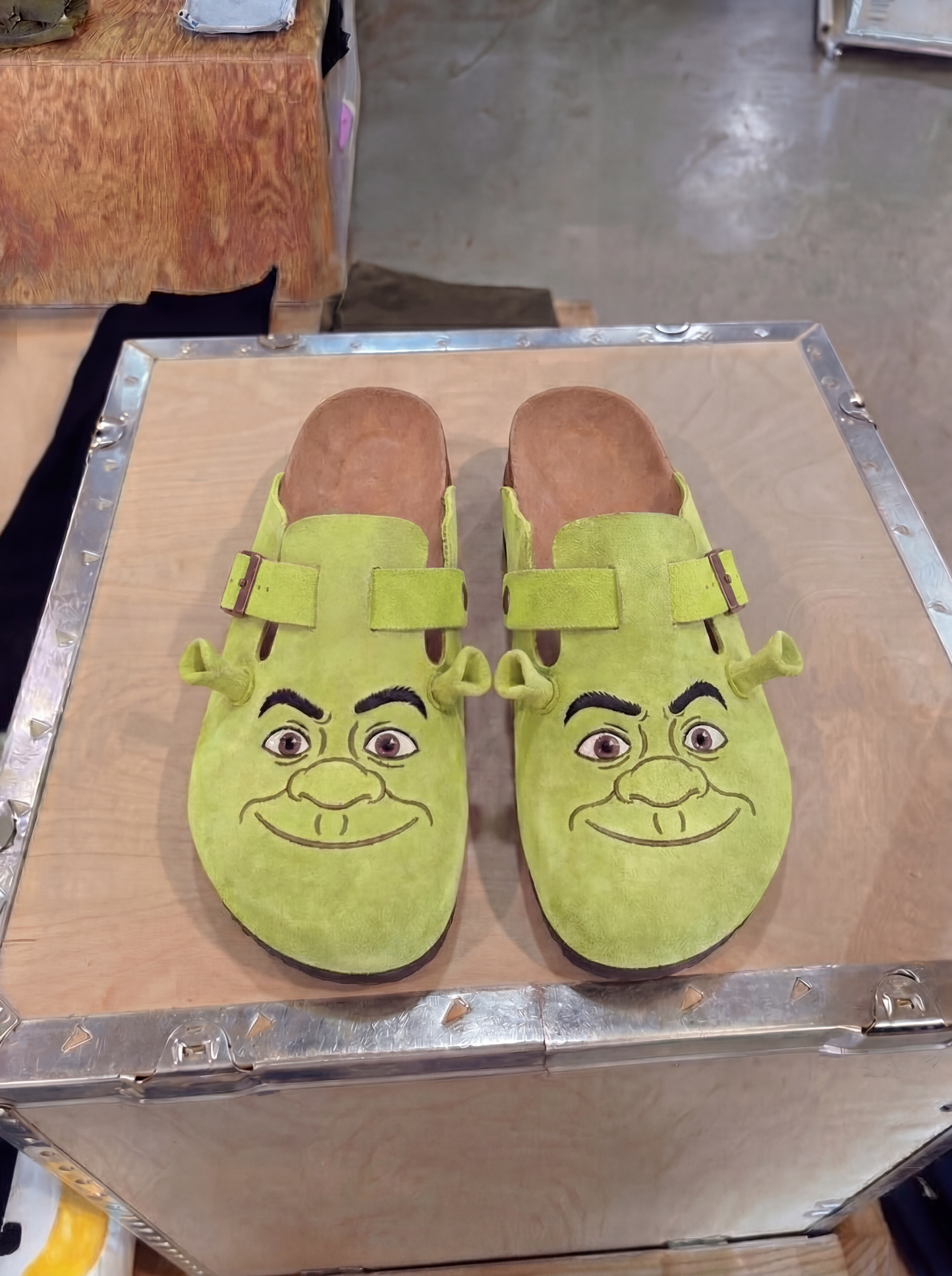 Pair of green Shrek-face suede clogs displayed on a wooden crate